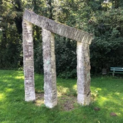 Anfang und Ende Steinskulptur mit zwei schrägen Säulen und Querbalken, umgeben von Bäumen und grünem Rasen.
