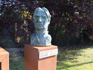 Skulptur Gottlieb Pot d'Or Gottlieb Pot d'Ors Büste steht auf einem metallenen Sockel und wird von Bäumen umrahmt.