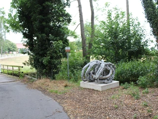 Skulptur Rolle vorwärts Skulptur Rolle vorwärts aus Stein, umgeben von Bäumen und einem asphaltierten Weg im Vordergrund.