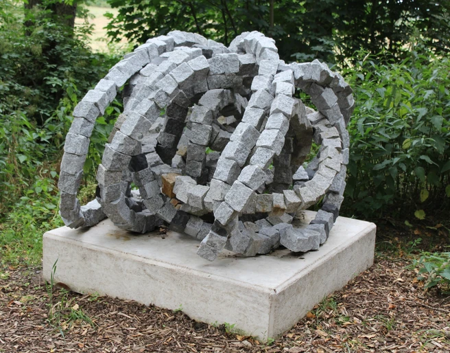 Skulptur Rolle vorwärts Steinskulptur "Rolle vorwärts" aus granitenen Segmentelementen auf Betonfundament, umgeben von Grün.