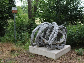 Skulptur Rolle vorwärts Skulptur aus verschlungenen Granitblöcken auf einem Podest, umgeben von üppigem Grün im Freien.