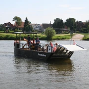 PetraSolara Personen auf einer kleinen Fähre überqueren einen ruhigen Fluss, umgeben von einer ländlichen Landschaft.