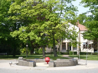 Alter Markt Kreisförmige Sitze mit zentraler rot-oranger Skulptur, umgeben von Bäumen und einem hellen Gebäude.