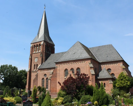 Kirche Lunsen Eine rote Backsteinkirche mit spitzem Turmdach steht vor einem klaren blauen Himmel in grüner Umgebung.