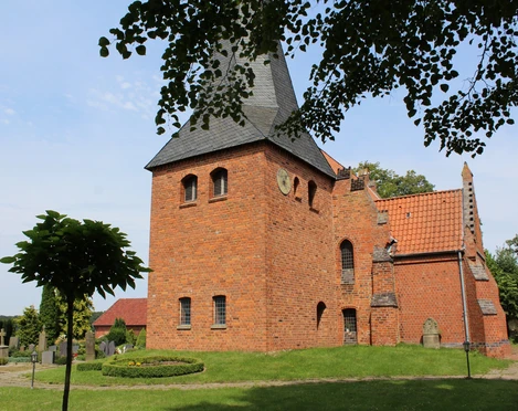 Die Kirche St. Crucis in Hoyerhagen aus rotem Backstein mit Turm steht vor blauem Himmel nahe Bäumen.