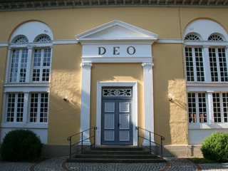 Gelbe Kirchenfassade mit weißen Säulen, der Schriftzug "DEO" und eine blaue Tür im Zentrum.