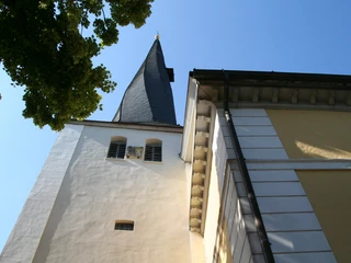Spitz zulaufender Kirchturm der St.-Jacobi-Kirche Stolzenau neben gelber Kirchenfassade unter klarem Himmel.
