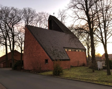 St. Marien Kirche Haßbergen Eine moderne Backsteinkirche mit steilem Dach, umgeben von Bäumen, aufgenommen in der Dämmerung.