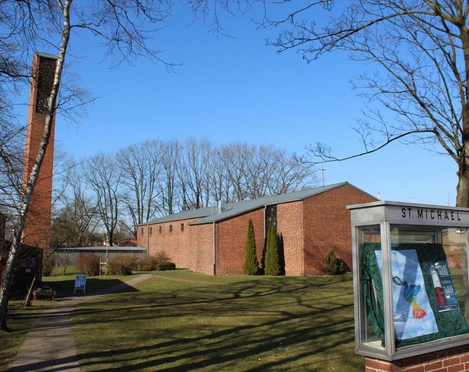 Backsteinkirche mit langem Kirchenschiff und freistehendem Glockenturm, umgeben von Rasen und Bäumen.