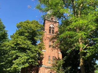 Backsteinkirchturm mit grünem Laub umgeben, unter strahlend blauem Himmel; enthält gotische Elemente.