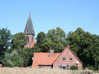 Backsteinkirche mit spitzem Turm hinter einem roten Ziegelhaus umgeben von Bäumen und Feldern.