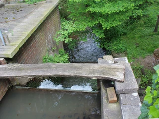 Wassermühle Heuermann Wassermühle mit hölzerner Brücke, Bach fließt zwischen Ziegelmauern und dichtem, grünem Blattwerk.