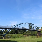 Wesertorbrücke Eine geschwungene blaue Fußgängerbrücke überspannt einen Fluss, umgeben von grüner Vegetation.