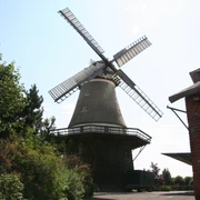 Windmühle Dörverden Windmühle mit weiß-roten Flügeln in ländlicher Umgebung, umgeben von Bäumen und einem Backsteingebäude.