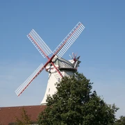 Windmühle mit roten Flügeln und weißen Holzplanken hinter Bäumen vor blauem Himmel in Langwedel-Etelsen.
