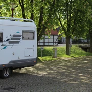 Reisemobillstellplatz Uchte Wohnmobil steht auf einem gepflasterten Platz, umgeben von grünen Bäumen und traditioneller Architektur.