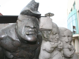 Skulptur eines Zigarrenmachers mit markantem Gesicht, flankiert von zwei neugierig beobachtenden Figuren.