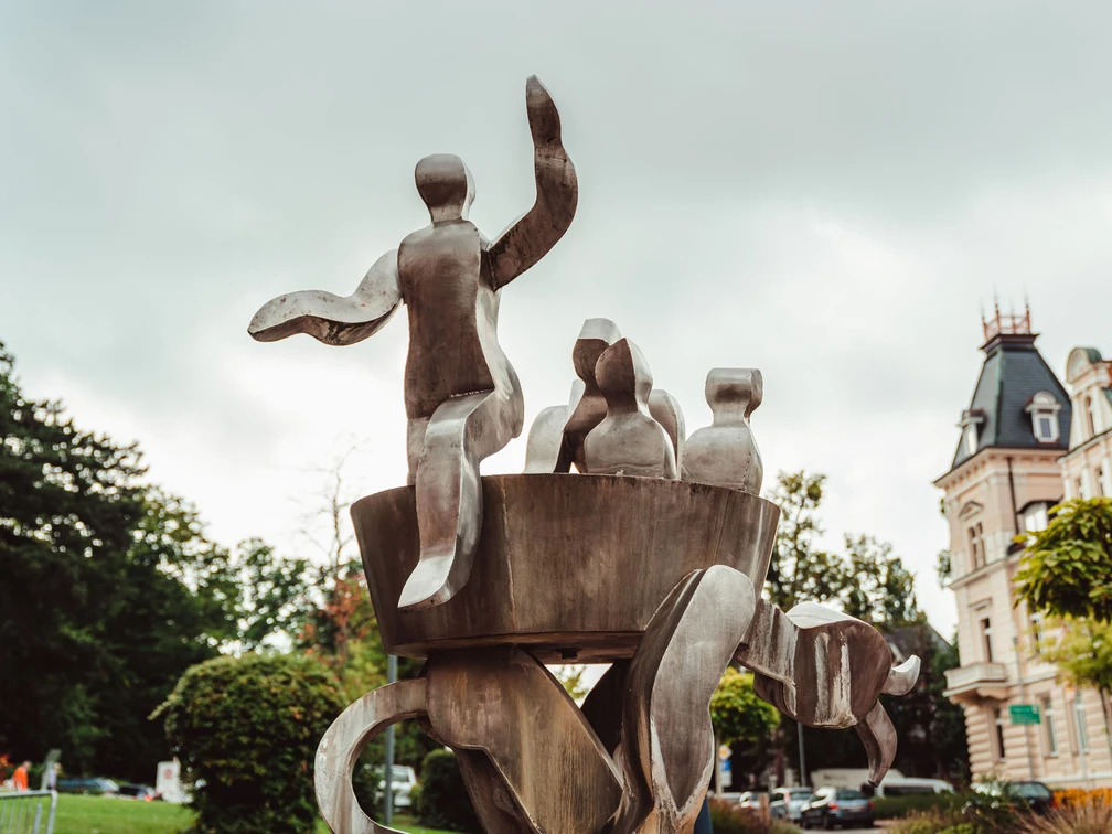 Brunnen "Auf dem Düvelsdreck" Metallskulptur mit abstrakten Figuren, umgeben von Bäumen und Gebäuden im Zentrum von Bad Nenndorf.