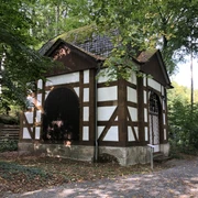 Friedrichspavillon.jpg Fachwerkpavillon im Grünen mit steinerner Basis und zwei großen, dunkelbraunen Holztüren.