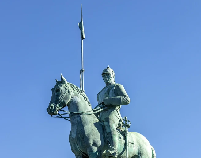 Cuirassier monument Bronzestatue eines Kürassiers zu Pferd auf einem hohen Sockel, mit einer Lanze über dem blauen Himmel. Bronze statue of a cuirassier on horseback on a high pedestal, with a lance above the blue sky.
