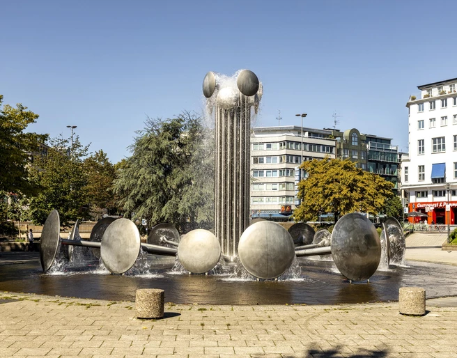 Ebertplatz Der Ebertplatz in Köln zeigt einen modernen Brunnen mit Wassertrommeln und einer zentralen Säule. Umgeben von Bäumen und Gebäuden, bietet er eine urbane Kulisse an einem sonnigen Tag. Ebertplatz in Cologne features a modern fountain with water drums and a central column. Surrounded by trees and buildings, it provides an urban backdrop on a sunny day.