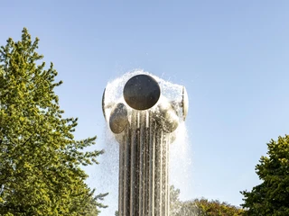 Ebertplatz Im Zentrum des Bildes erhebt sich eine große, futuristische Skulptur mit Wasserfontänen. Umgeben von kugelförmigen Elementen, schimmert der Brunnen im Sonnenlicht, eingerahmt von üppigem Grün der umliegenden Bäume.A large, futuristic sculpture with water fountains rises in the center of the picture. Surrounded by spherical elements, the fountain shimmers in the sunlight, framed by the lush green of the surrounding trees.