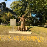 Die Melkerin Eine Bronzeskulptur einer Frau mit einem Joch und Milchkannen steht vor einer Infotafel in einem Park.