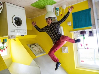 das-verrueckte-haus.jpg Das Bild zeigt einen Mann, der freudig in einem Raum steht, dessen Einrichtung kopfüber an der Decke angebracht ist. Zu sehen sind eine Waschmaschine, ein WC und bunte Wände in einem surrealen, umgekehrten Haus.