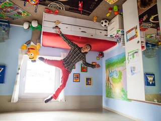 das-verrueckte-haus-balanceakt (1).jpg Ein Mann steht auf der Decke eines bunt gestalteten Kinderzimmers, als befände er sich in einem verkehrten Raum.