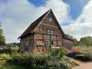 Noltesche Scheune Eine historische Fachwerkscheune, umgeben von grünen Büschen und Blumen, unter einem teils bewölkten Himmel.