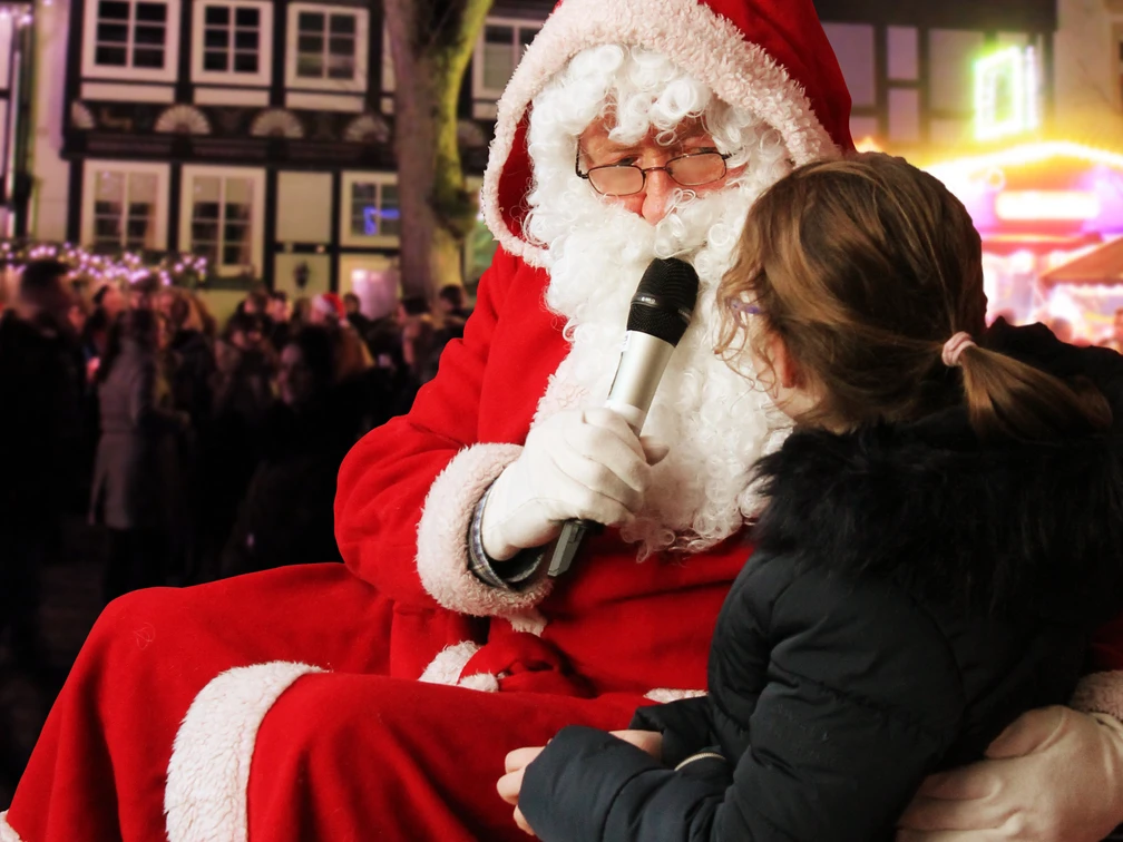 1.jpg Weihnachtsmann spricht lächelnd mit einem Kind, im Hintergrund festlich geschmückte Fachwerkhäuser.