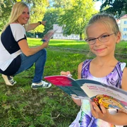 Stadtspiel Sebnitz Mädchen mit Brille liest ein buntes Buch im Park, während eine Frau im Hintergrund lächelnd auf etwas zeigt.