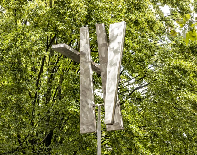 Four Trapezoids as two Rectangles IV Eine moderne Skulptur aus Aluminium-Trapezen erhebt sich vor dichtem grünen Laub.A modern sculpture made of aluminum trapezoids rises up in front of dense green foliage.