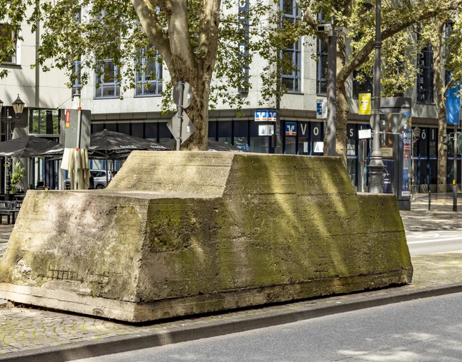 Motionless traffic Ein moosbedeckter Betonblock mit Bäumen darauf steht inmitten einer urbanen Straßenszene.A moss-covered concrete block with trees on it stands in the middle of an urban street scene.