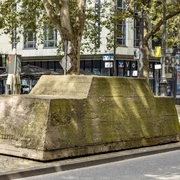 Ruhender Verkehr Ein moosbedeckter Betonblock mit Bäumen darauf steht inmitten einer urbanen Straßenszene.A moss-covered concrete block with trees on it stands in the middle of an urban street scene.