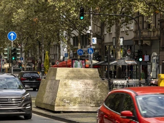 Motionless traffic Am Bild sind parkende und fahrende Autos in einer belebten Kölner Straße zu sehen, flankiert von Geschäften und Bäumen. Ein massiver Betonblock in der Straßenmitte hebt sich inmitten des urbanen Verkehrs hervor, während die Ampeln den fließenden Verkehr steuern.The picture shows parked and moving cars in a busy street in Cologne, flanked by stores and trees. A massive concrete block in the middle of the street stands out amidst the urban traffic, while the traffic lights control the flow of traffic.