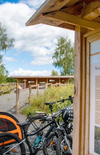 Neustart in der Tongrube.jpg Fahrräder und ein Hundewagen vor einer Info-Tafel in einer landschaftlichen Naturanlage.