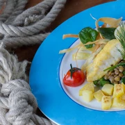 Bio-Restaurant StrandGut Ein Teller mit paniertem Fisch, Kartoffelwürfeln, Linsen, Spinat, Tomaten und Zitronenscheibe auf blauem Rand, neben einem Seil auf Holztisch.