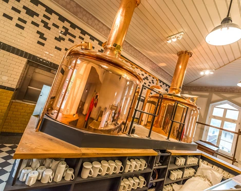 Kupferne Braukessel in einer Brauerei mit weißen und schwarzen Fliesen an den Wänden; darunter Regale mit weißen Krügen.