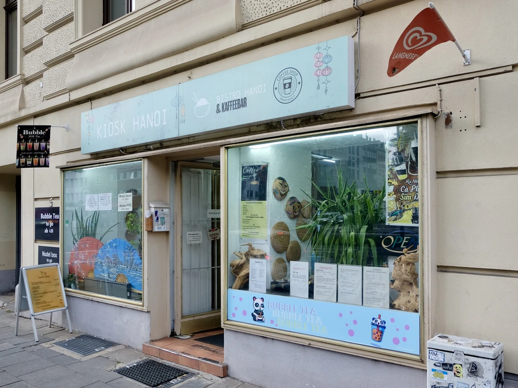 Kiosk Hanoi Die Fassade ist mit großen Fenstern ausgestattet, hinter denen Dekorationen und Pflanzen zu sehen sind. Ein Schild mit dem Namen des Geschäfts und Informationen über das angebotene Sortiment befindet sich oberhalb der Fenster. Vor dem Kiosk steht ein Aufsteller mit weiteren Details zu den angebotenen Produkten. Die Fenster sind mit Illustrationen von Bubble Tea verziert.The façade has large windows, behind which decorations and plants can be seen. A sign with the name of the store and information about the range on offer is located above the windows. In front of the kiosk is a display with further details about the products on offer. The windows are decorated with illustrations of bubble tea.