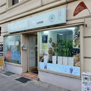 Kiosk Hanoi Die Fassade ist mit großen Fenstern ausgestattet, hinter denen Dekorationen und Pflanzen zu sehen sind. Ein Schild mit dem Namen des Geschäfts und Informationen über das angebotene Sortiment befindet sich oberhalb der Fenster. Vor dem Kiosk steht ein Aufsteller mit weiteren Details zu den angebotenen Produkten. Die Fenster sind mit Illustrationen von Bubble Tea verziert.The façade has large windows, behind which decorations and plants can be seen. A sign with the name of the store and information about the range on offer is located above the windows. In front of the kiosk is a display with further details about the products on offer. The windows are decorated with illustrations of bubble tea.