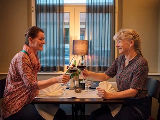 Logis Hotel Quartier 5 Zwei Frauen sitzen an einem Tisch in einem Restaurant und stoßen mit Rotweingläsern an, lächelnd und in entspannter Atmosphäre.