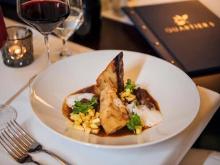 Logis Hotel Quartier 5 Ein elegantes Gericht mit Fleisch, Schaum und Gemüse auf weißem Teller, umgeben von Weingläsern und einer Kerze auf einem gedeckten Tisch.