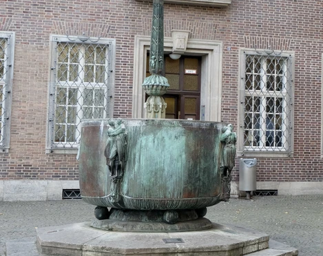 Carnival fountain Der Fastnachtsbrunnen in Köln zeigt eine künstlerische Skulptur mit Bronzefiguren, die die berühmte fünfte Jahreszeit darstellt. Der Brunnen ist ein Kunstwerk mit detaillierten Figuren, das auf einem gepflasterten Vorplatz vor einem Backsteingebäude steht. Eine Säule in der Mitte, gekrönt mit symbolischen Karnevalsfiguren, zieht den Blick auf sich. Umrahmt von sechs Bronzemasken, steht es stellvertretend für Fröhlichkeit und Tradition des rheinischen Karnevals.The Fastnachtsbrunnen in Cologne features an artistic sculpture with bronze figures depicting the famous fifth season. The fountain is a work of art with detailed figures that stands on a paved forecourt in front of a brick building. A column in the middle, crowned with symbolic carnival figures, draws the eye. Framed by six bronze masks, it represents the cheerfulness and tradition of the Rhineland carnival.