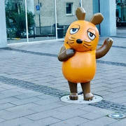 Mouse statue Die Maus-Statue mit erhobenem Arm, ein Symbol der beliebten Kindersendung, steht auf Gehwegplatten vor einem modernen Gebäude in Köln.The mouse statue with raised arm, a symbol of the popular children's show, stands on paving slabs in front of a modern building in Cologne.