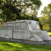 Monument to the gray buses Das Denkmal der grauen Busse zeigt ein stilisiertes, lebensgroßes Betonabbild eines Busses, symbolträchtig in einem Park platziert. Umgeben von Bäumen, fügt sich die Skulptur harmonisch in die grüne Umgebung ein.The monument to the grey buses shows a stylized, life-size concrete image of a bus, symbolically placed in a park. Surrounded by trees, the sculpture blends harmoniously into the green surroundings.