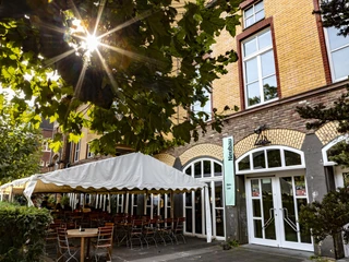 Alte Feuerwache Die Alte Feuerwache hat eine historische Fassade aus gelben Ziegeln mit hoher Fensterfront. Ein Sonnenschirm beschattet zahlreiche Außenplätze, eingerahmt von grünen Pflanzen.The Alte Feuerwache has a historic façade made of yellow bricks with a high window front. A parasol shades numerous outdoor seats, framed by green plants.