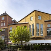 Alte Feuerwache Alte Feuerwache Köln mit einer markanten Kombination aus Backsteinfassaden und modernen Holzelementen in warmen Gelbtönen. Im Vordergrund stehen Bäume, die dem Gebäude eine grüne Umrahmung verleihen.Old Cologne fire station with a striking combination of brick façades and modern wooden elements in warm yellow tones. In the foreground are trees that give the building a green frame.