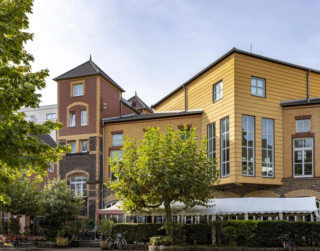 Alte Feuerwache Alte Feuerwache Köln mit einer markanten Kombination aus Backsteinfassaden und modernen Holzelementen in warmen Gelbtönen. Im Vordergrund stehen Bäume, die dem Gebäude eine grüne Umrahmung verleihen.Old Cologne fire station with a striking combination of brick façades and modern wooden elements in warm yellow tones. In the foreground are trees that give the building a green frame.