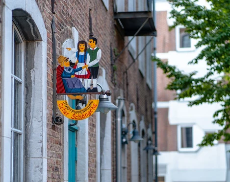Buntes Puppentheater-Schild „Öcher Schängche“ an einer Backsteinfassade. Zwei stilisierte Puppen sind darauf abgebildet.